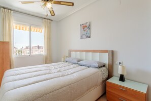 Ein Schlafzimmer mit Bett, Nachttisch, Lampe, Deckenventilator, einem Fenster mit Vorhängen und einem Wandbild.