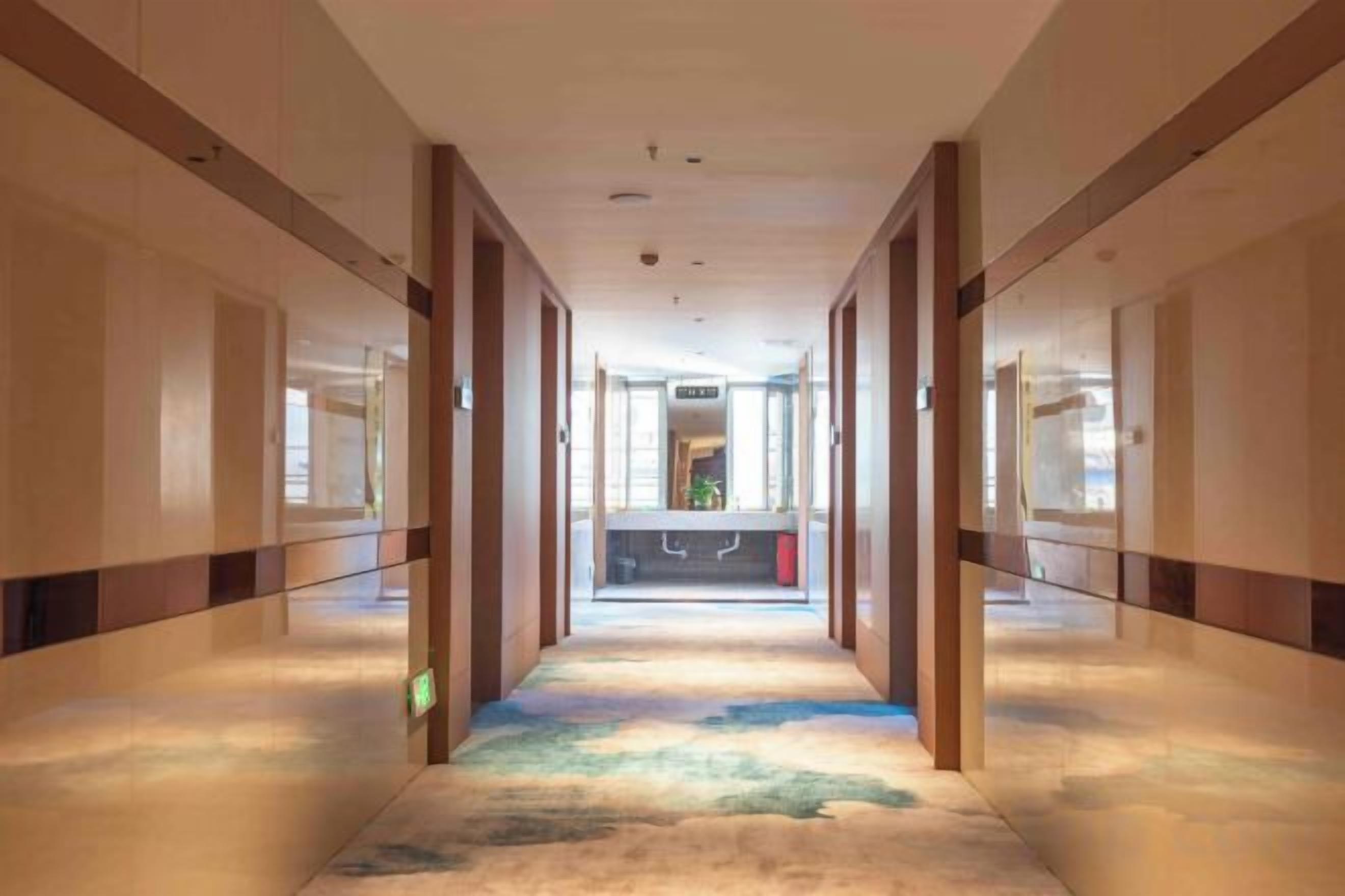 A hotel corridor with a carpeted floor, doors on both sides, and a reception area in the distance.