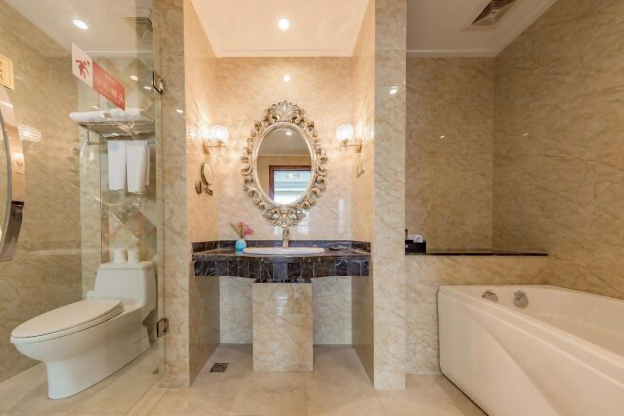 Un baño con pared de mármol, inodoro blanco, lavamanos con espejo y bañera.