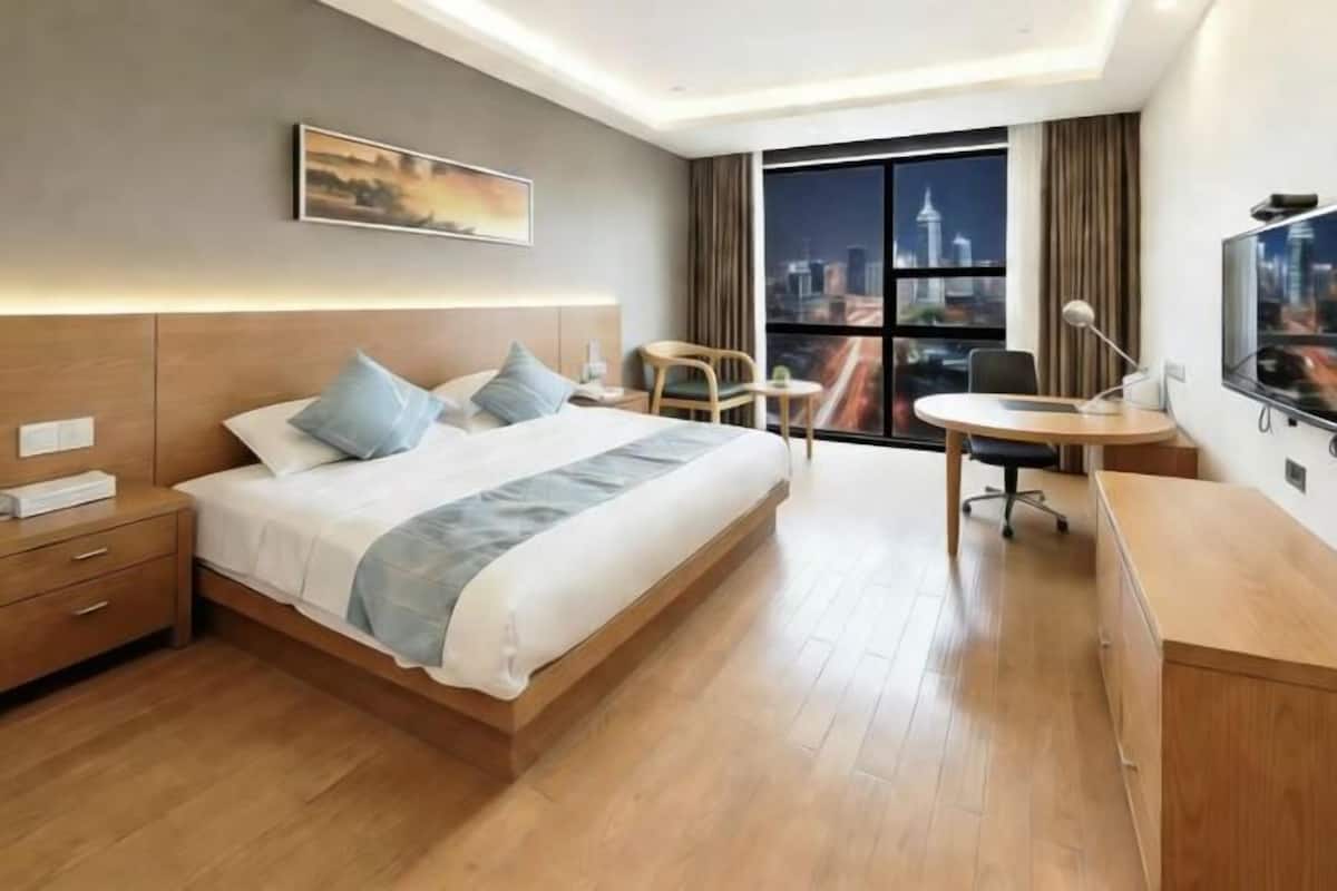 Una habitación de hotel moderna con una cama grande, un escritorio, una silla y vistas a un paisaje urbano a través de grandes ventanales.