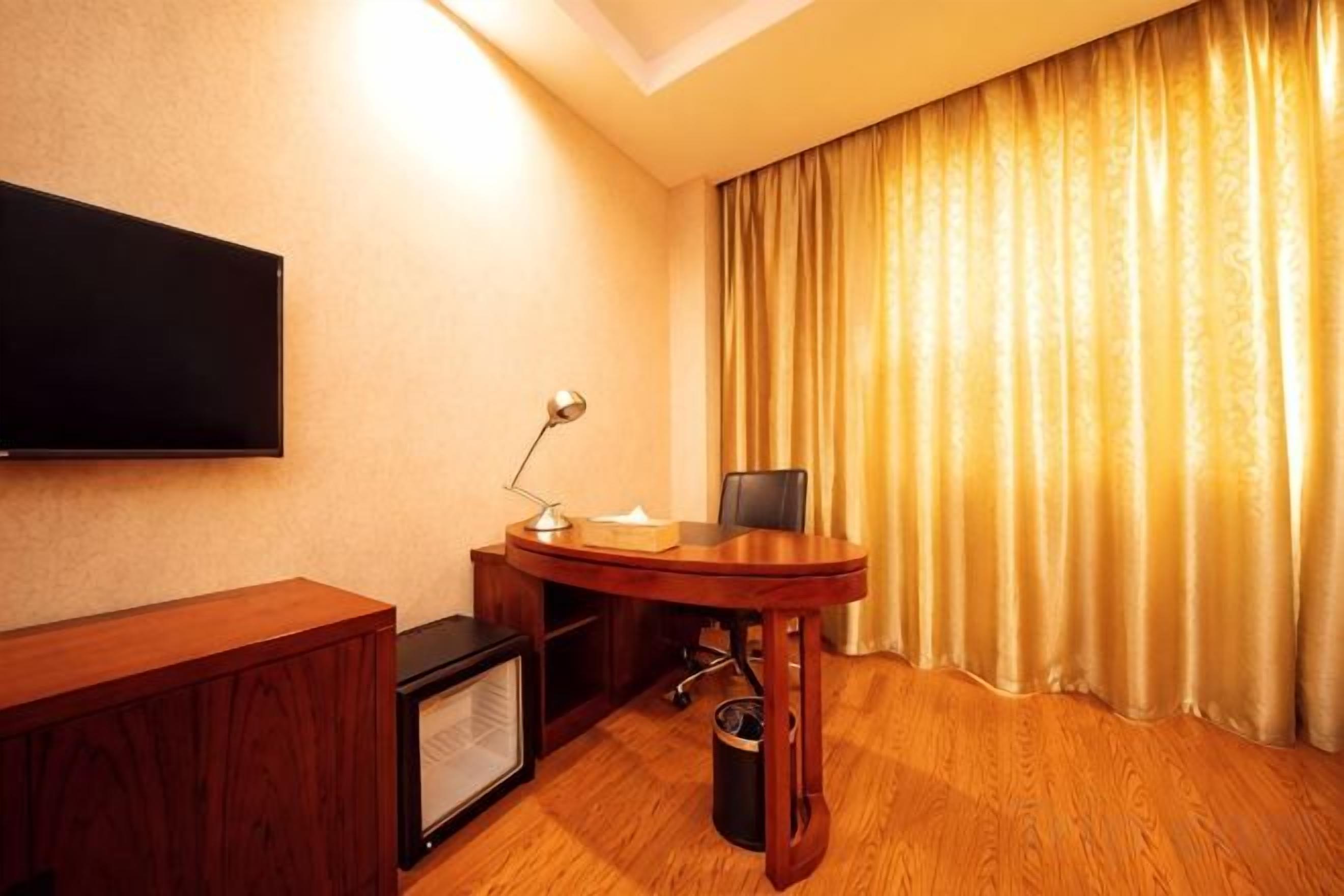 A hotel room with a flat-screen TV mounted on the wall, a wooden desk with a chair, a small table with a lamp, and a large window with curtains.