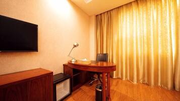 A hotel room with a flat-screen TV mounted on the wall, a wooden desk with a chair, a small table with a lamp, and a large window with curtains.