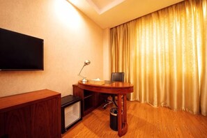 Une chambre d’hôtel équipée d’un téléviseur à écran plat fixé au mur, d’un bureau en bois avec une chaise, d’une petite table avec une lampe et d’une grande fenêtre avec des rideaux.