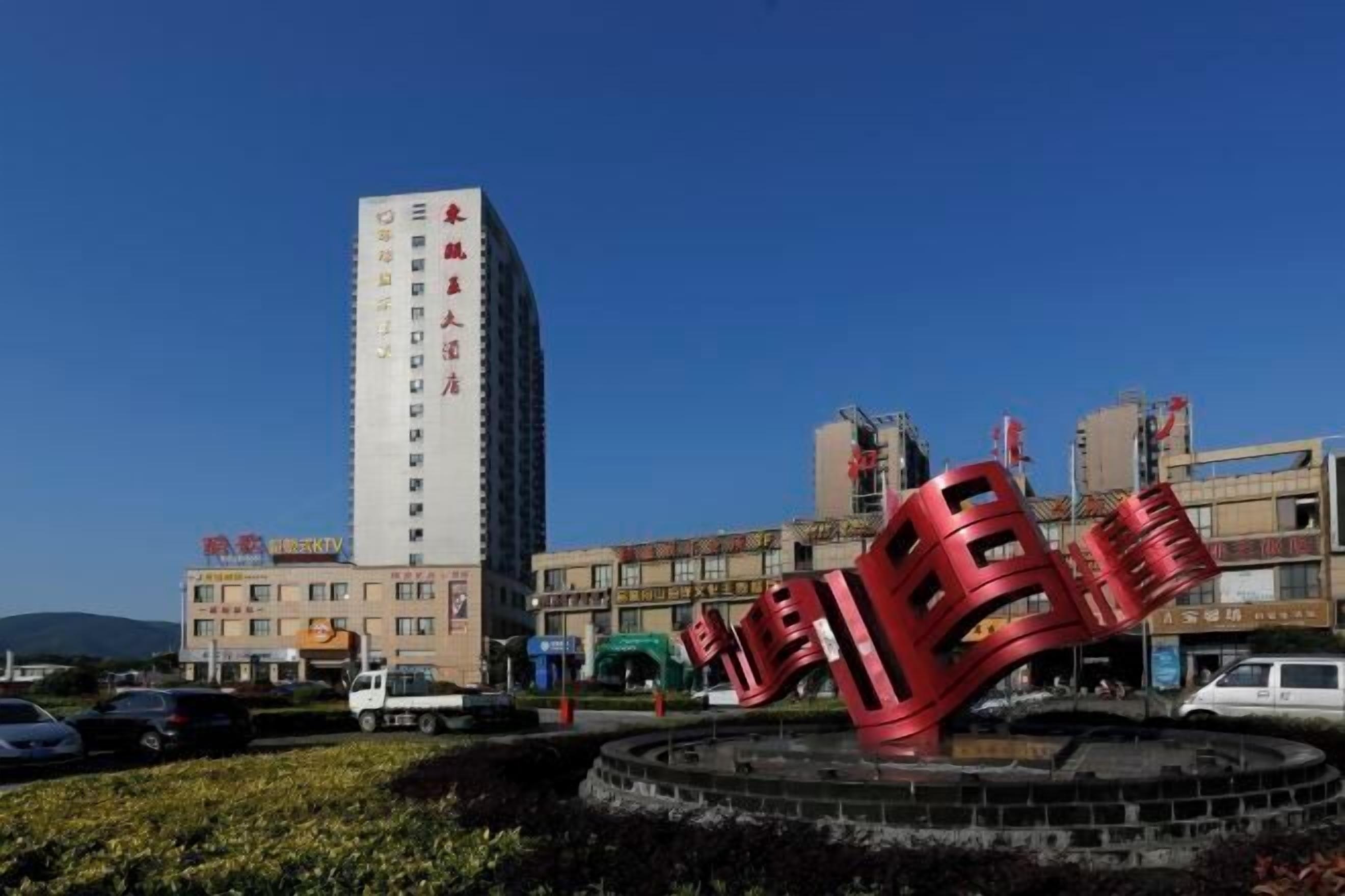 Ein modernes Gebäude mit vertikaler Gestaltung, ein kreisförmiger Platz mit einer roten Skulptur und einem strahlend blauen Himmel.
