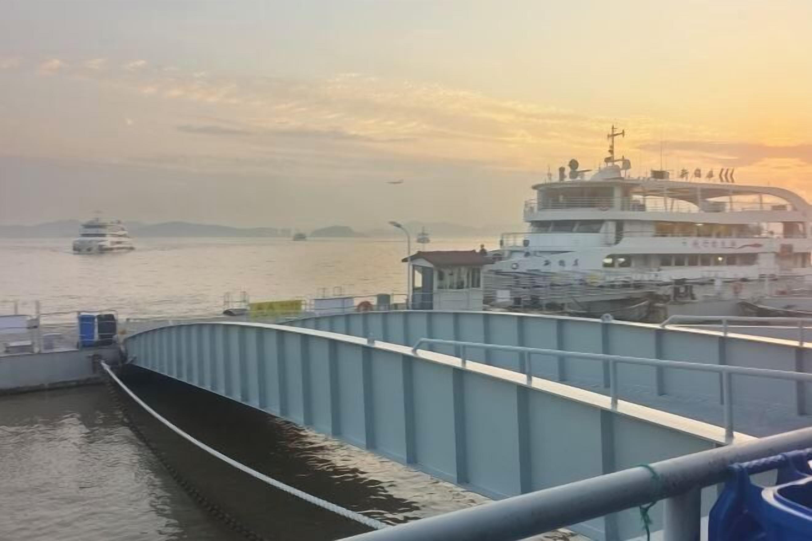 Ein Fährterminal mit einer großen Fähre im Hafen, eine Brücke im Vordergrund und ein Sonnenuntergang im Hintergrund.