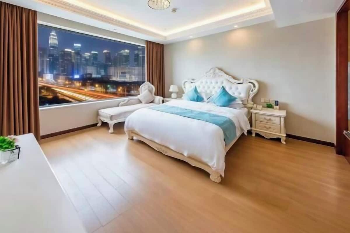 Una habitación de hotel moderna con una cama grande, una mesita de noche y vistas a un paisaje urbano a través de un ventanal amplio.