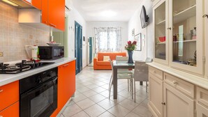 Een moderne keuken met een gasfornuis, magnetron en oranje kastjes. Er is een eethoek met een tafel en stoelen, en een woonkamer met een bank en een raam.