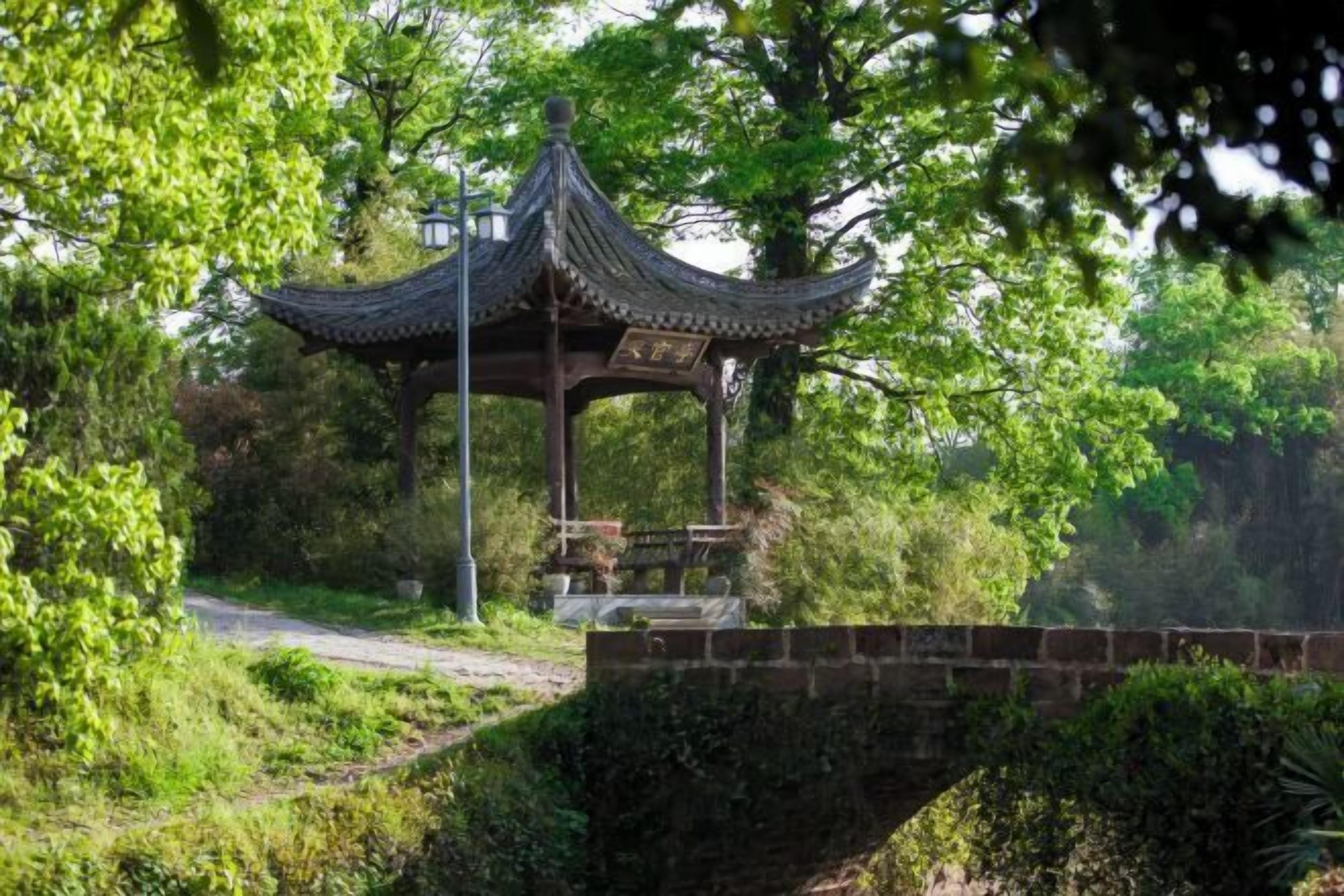 A traditional pavilion with a tiled roof, surrounded by lush greenery and a stone bridge.