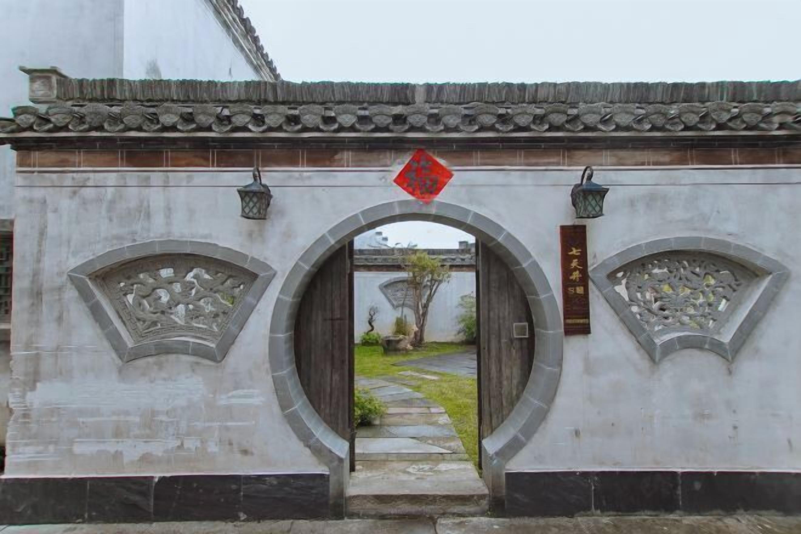 Une porte chinoise traditionnelle, ornée de sculptures complexes, surmontée d’un toit en tuiles et bordée d’un sentier qui la traverse.