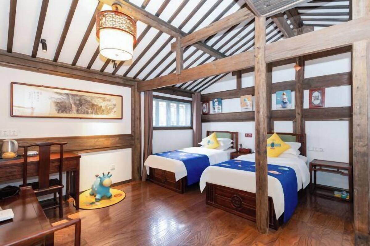 Quarto com vigas de madeira, duas camas com roupa de cama azul e branca, uma escrivaninha de madeira, uma cadeira, uma mesinha e uma luminária, além de um quadro na parede.