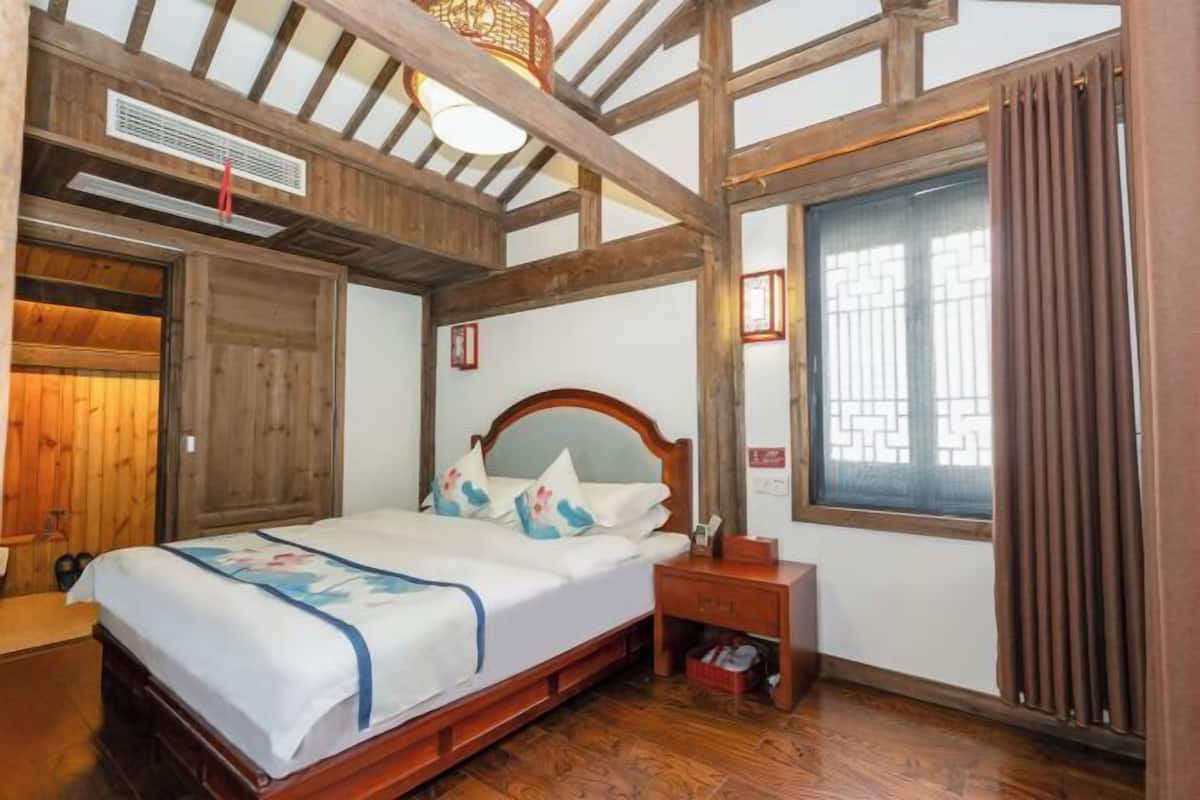 Quarto com cama de madeira, mesa de cabeceira, janela com cortinas e estrutura de teto em madeira.