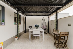 En overdækket terrasse med et hvidt bord og stole, en hængestol og potteplanter.