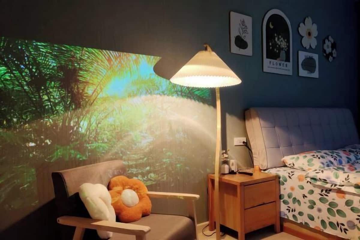 Une chambre à coucher comprenant un lit, une table de chevet, une lampe, une chaise et un projecteur mural.