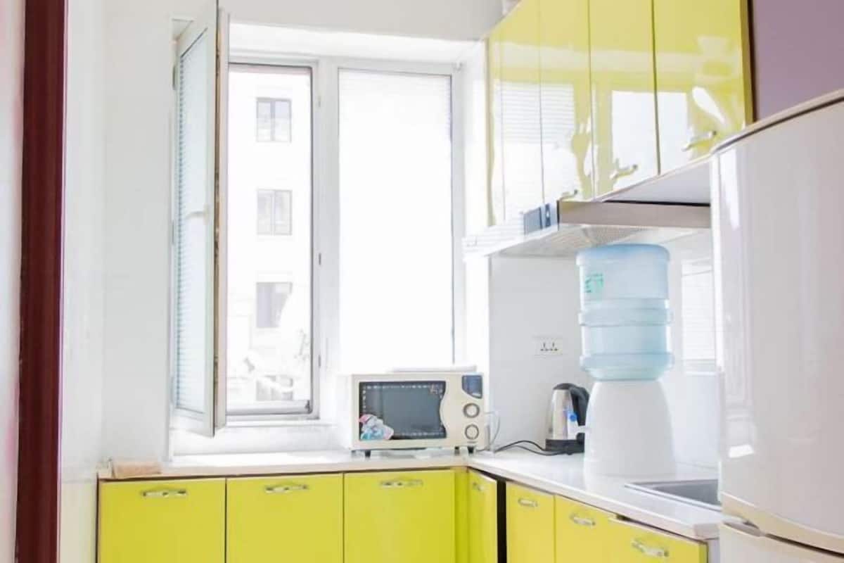 Une cuisine avec des meubles de couleur jaune, un four à micro-ondes, un distributeur d’eau et une fenêtre équipée de stores.