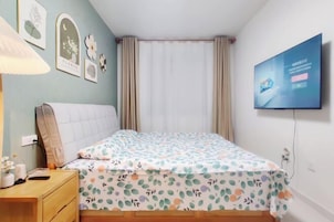 Un dormitorio con cama, mesita de noche, televisor montado en la pared y cortinas en la ventana.