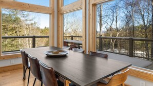 A dining area with a large wooden table and chairs, large windows, and a view of trees. - Modern Mountain Lodge with a View (Fleetwood)