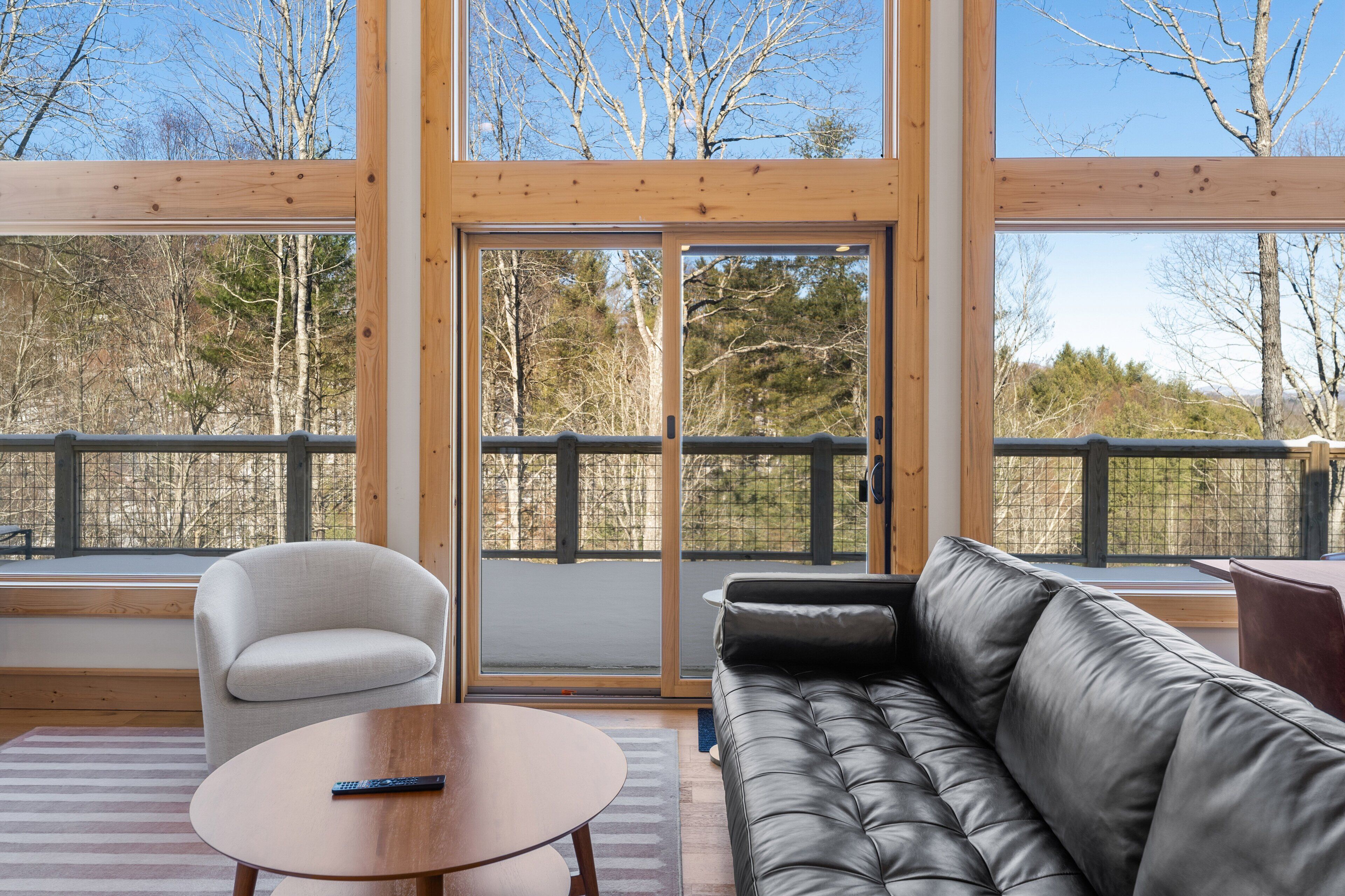 A modern room with a large sliding glass door leading to a balcony, a grey sofa, a round wooden table, and a light grey armchair.