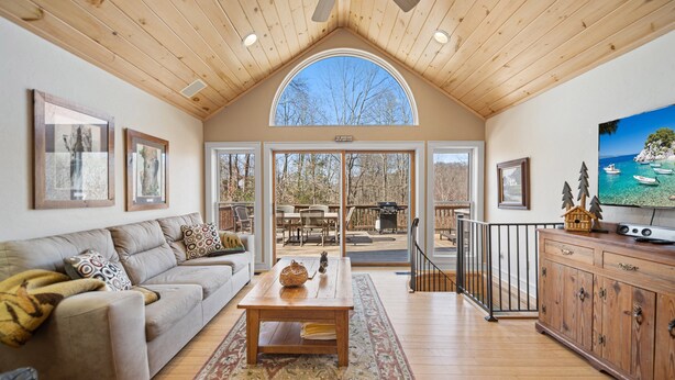 A spacious living room with a wooden ceiling, a large window, a television, and a dining area visible through glass doors. - Updated Mountain Home in Private Setting (Banner Elk)