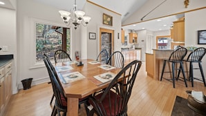 A spacious dining area with a wooden table, black chairs, and a chandelier. A kitchen with wooden cabinets and a countertop is visible in the background. - Updated Mountain Home in Private Setting (Banner Elk)