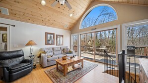 A spacious living area with a wooden ceiling, a leather sofa, a gray couch, and a wooden coffee table. Large windows provide a view of the outdoors. - Updated Mountain Home in Private Setting (Banner Elk)