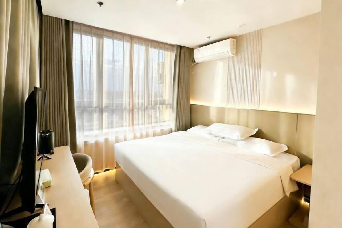 A hotel room with a large bed, a desk, a chair, an air conditioning unit, and a window with curtains.