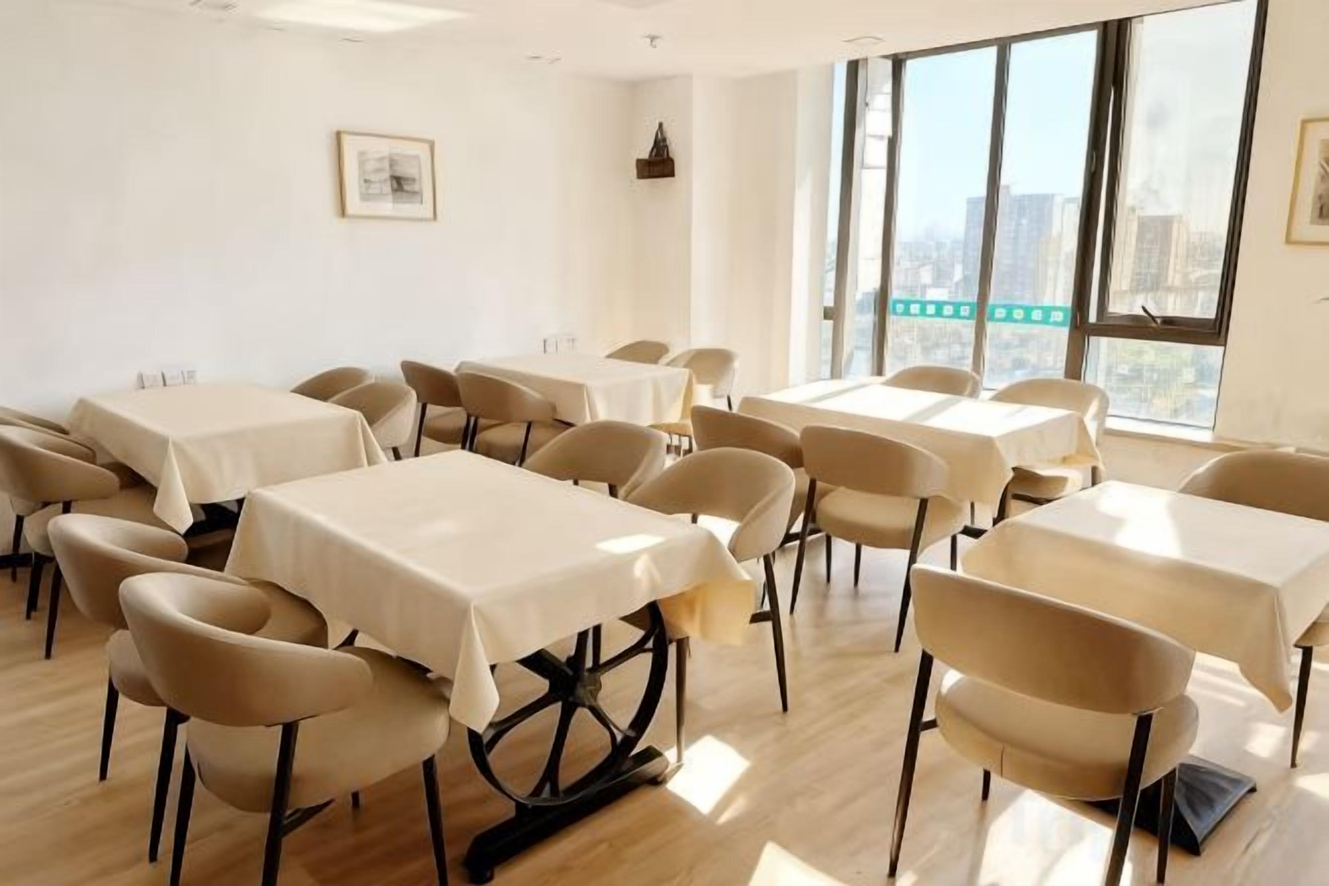 Une salle de conférence dotée de plusieurs tables et chaises, de grandes fenêtres offrant une vue sur les bâtiments et d’un tableau encadré accroché au mur.