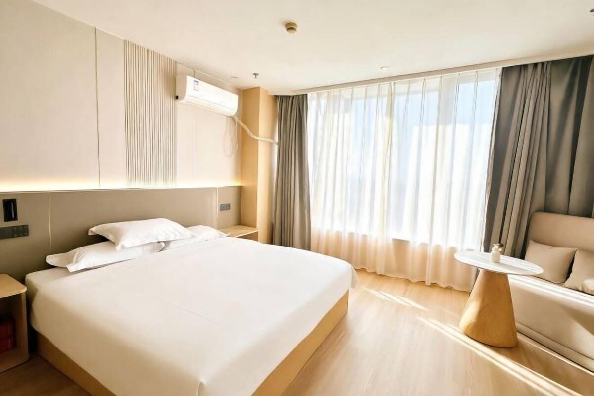 A hotel room with a large bed, a bedside table, a small round table, a sofa, and a window with curtains.