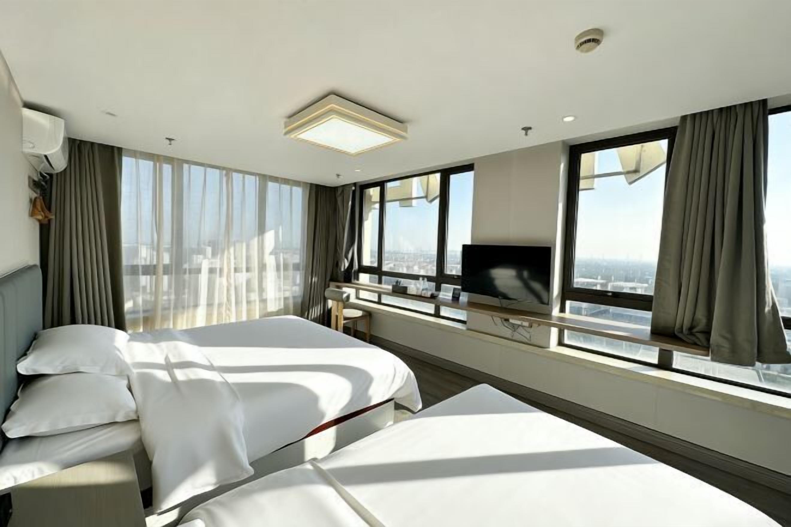 A hotel room with two beds, a TV, a desk, and large windows with curtains.