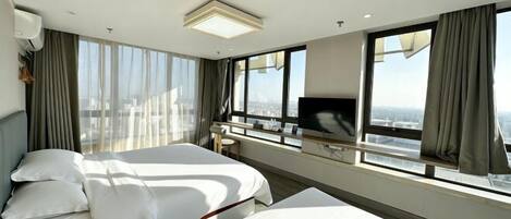 A hotel room with two beds, a TV, a desk, and large windows with curtains.