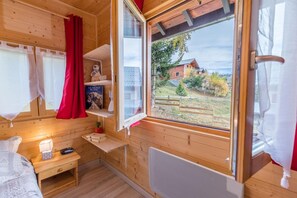 Ett mysigt rum i en stuga med träpanel på väggarna, ett fönster med utsikt över ett hus och träd, ett litet nattduksbord med en lampa och en hylla med dekorativa föremål.