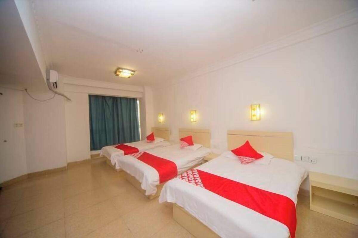 Quarto de hotel com três camas, cada uma com colcha vermelha e branca, cabeceira de madeira e mesa de cabeceira.
