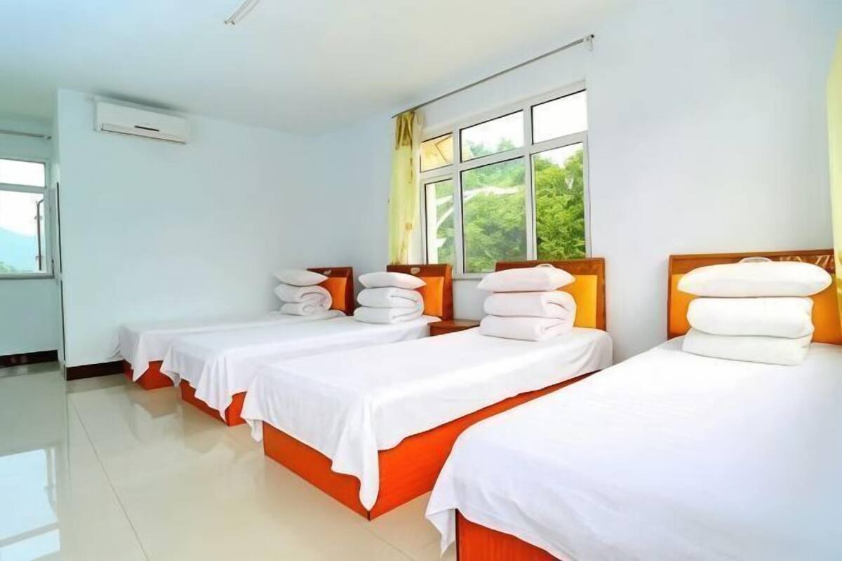 Quarto de hotel com três camas, cada uma com roupa de cama branca e cabeceiras laranja, janelas grandes com cortinas e chão de azulejos.
