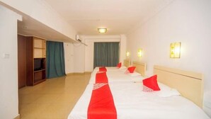 Habitación de hotel con tres camas alineadas, cada una con ropa de cama blanca y detalles rojos. Se aprecia un armario de madera y una cortina verde.