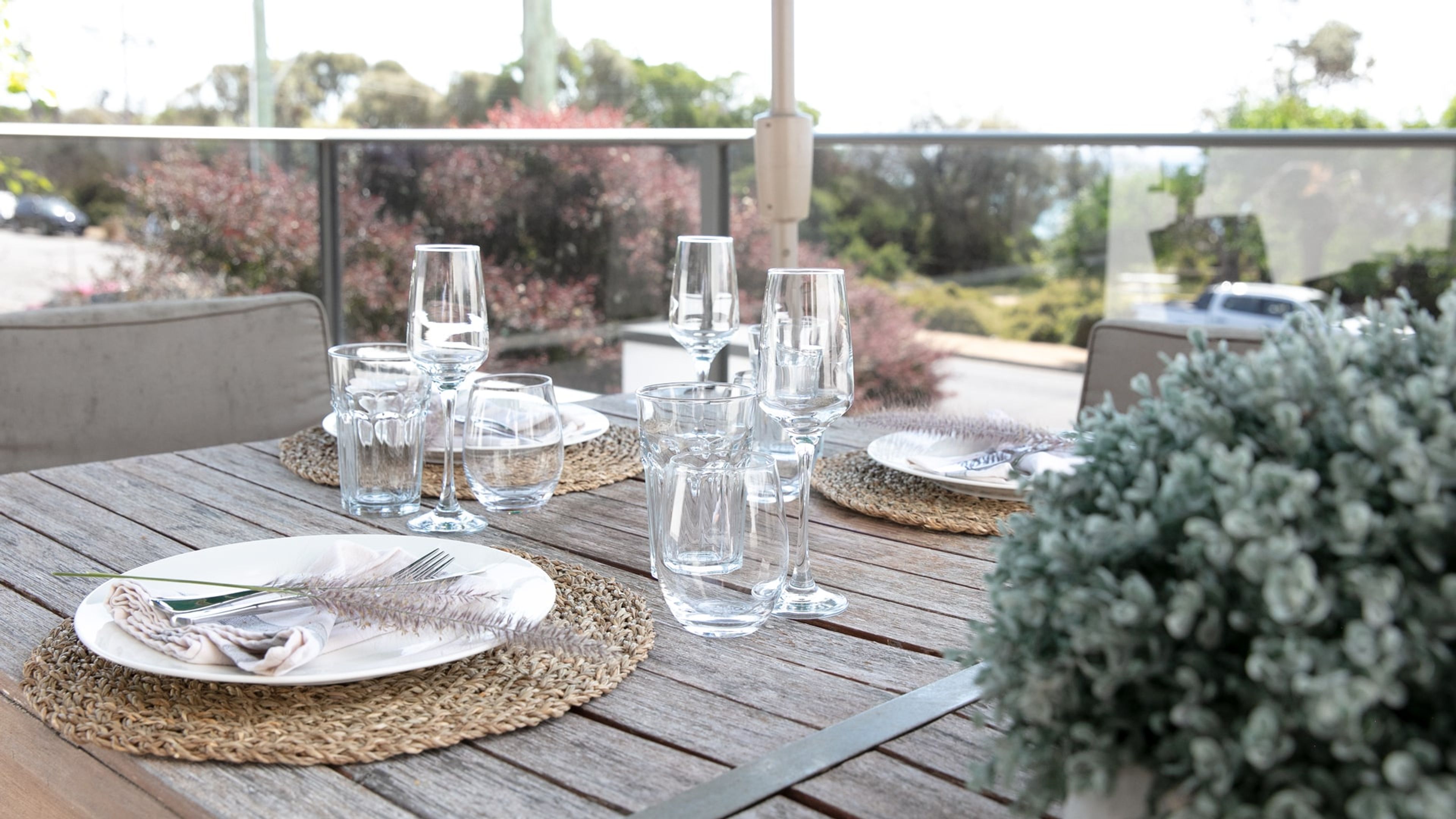 A wooden table set with clear glassware, plates, and cutlery, with a view of trees and a building in the background.