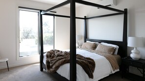 A four-poster bed with a fur throw, a bedside table with a lamp, and a view of the outdoors through large windows. - Stunning Beach Views Pet Friendly Dromana Retreat (Dromana)