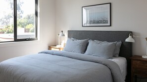 Un dormitorio con cama, mesita de noche, lámpara, un cuadro en la pared y una ventana con vista a árboles.