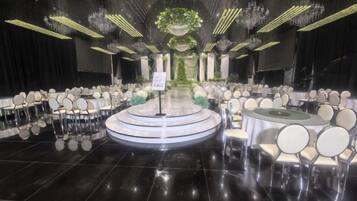 Un salón amplio y bien iluminado con filas de sillas y mesas, un escenario central y una araña de cristal.