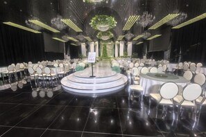 Um salão amplo e bem iluminado, com fileiras de cadeiras e mesas, palco central e um lustre.