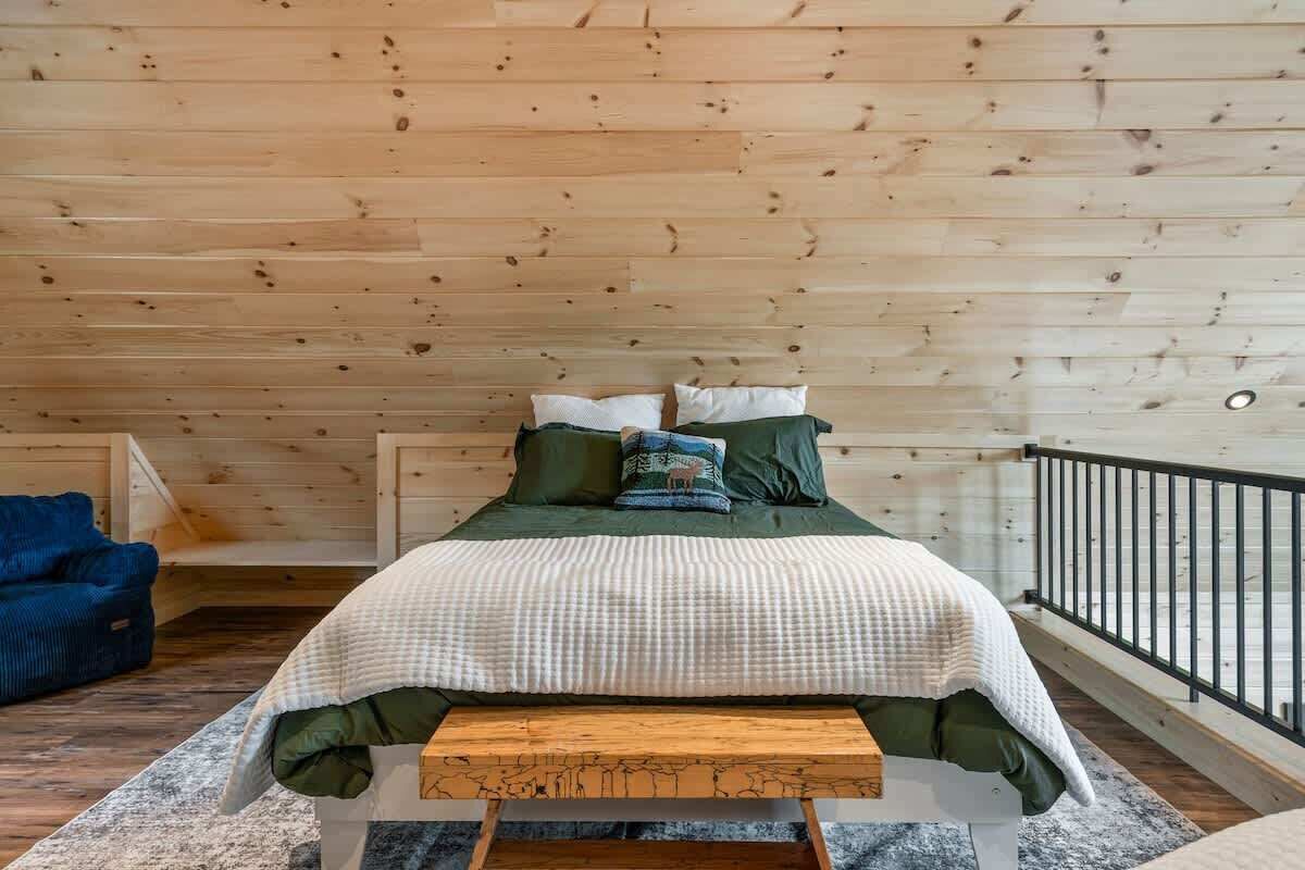 A bedroom with a wooden ceiling, a bed with green bedding, a blue chair, and a wooden bench.
