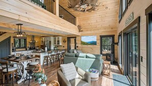 Un interior espacioso con techo alto, una zona de estar central, una cocina y un ventanal con vistas al exterior.
