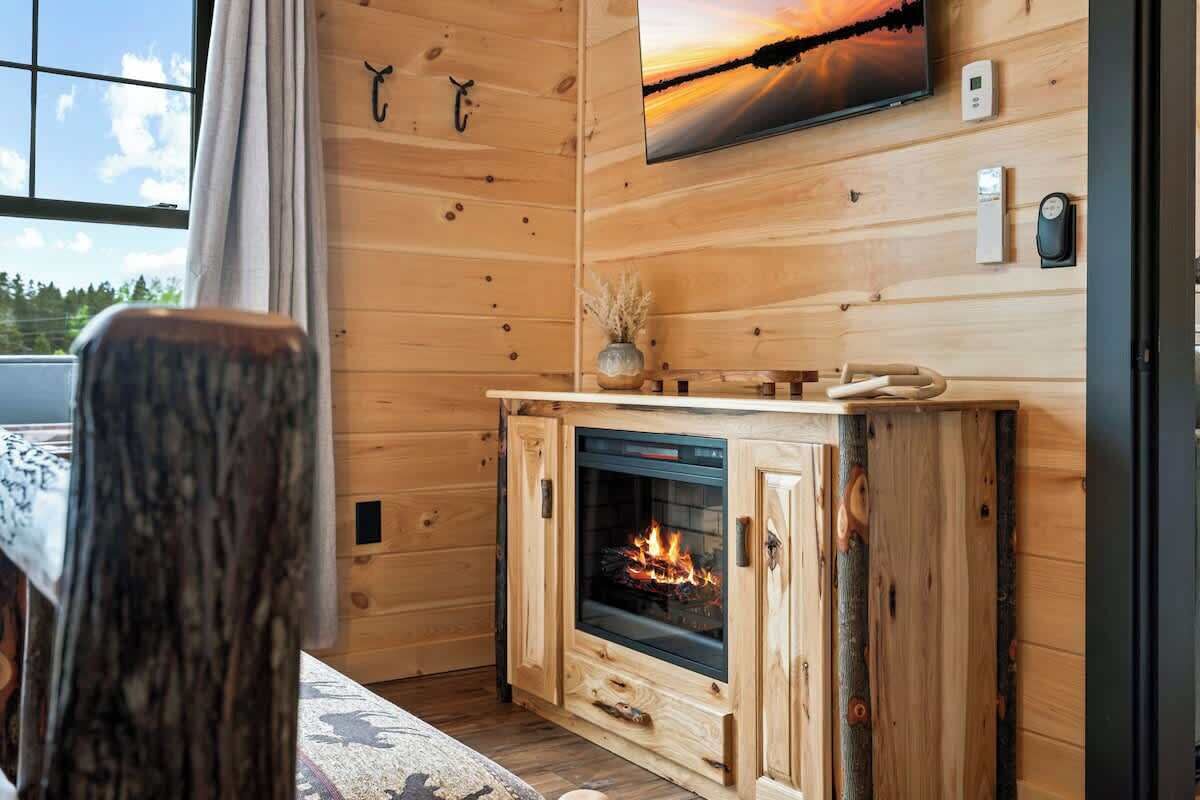 Ein gemütliches Blockhaus-Interieur mit Holzvertäfelung, Kamin, Wand-Fernseher und Fenster mit Blick ins Freie.