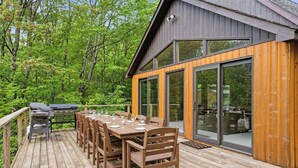 Une terrasse en bois avec une table et des chaises pour les repas, un barbecue et un bâtiment entièrement vitré doté de grandes fenêtres.