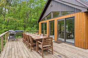 Une terrasse en bois avec une table et des chaises pour les repas, un barbecue et un bâtiment entièrement vitré doté de grandes fenêtres.