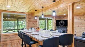 Un intérieur en bois avec une longue table à manger dressée pour quatre personnes, une cheminée et une grande fenêtre donnant sur un paysage verdoyant.
