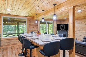 Un intérieur en bois avec une longue table à manger dressée pour quatre personnes, une cheminée et une grande fenêtre donnant sur un paysage verdoyant.