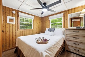 Un dormitorio con paneles de madera, una cama, un ventilador de techo, un aparador y una ventana con vistas a la vegetación.