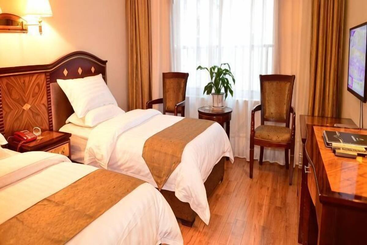 A hotel room with two beds, a desk, a chair, a lamp, a plant, and a television.