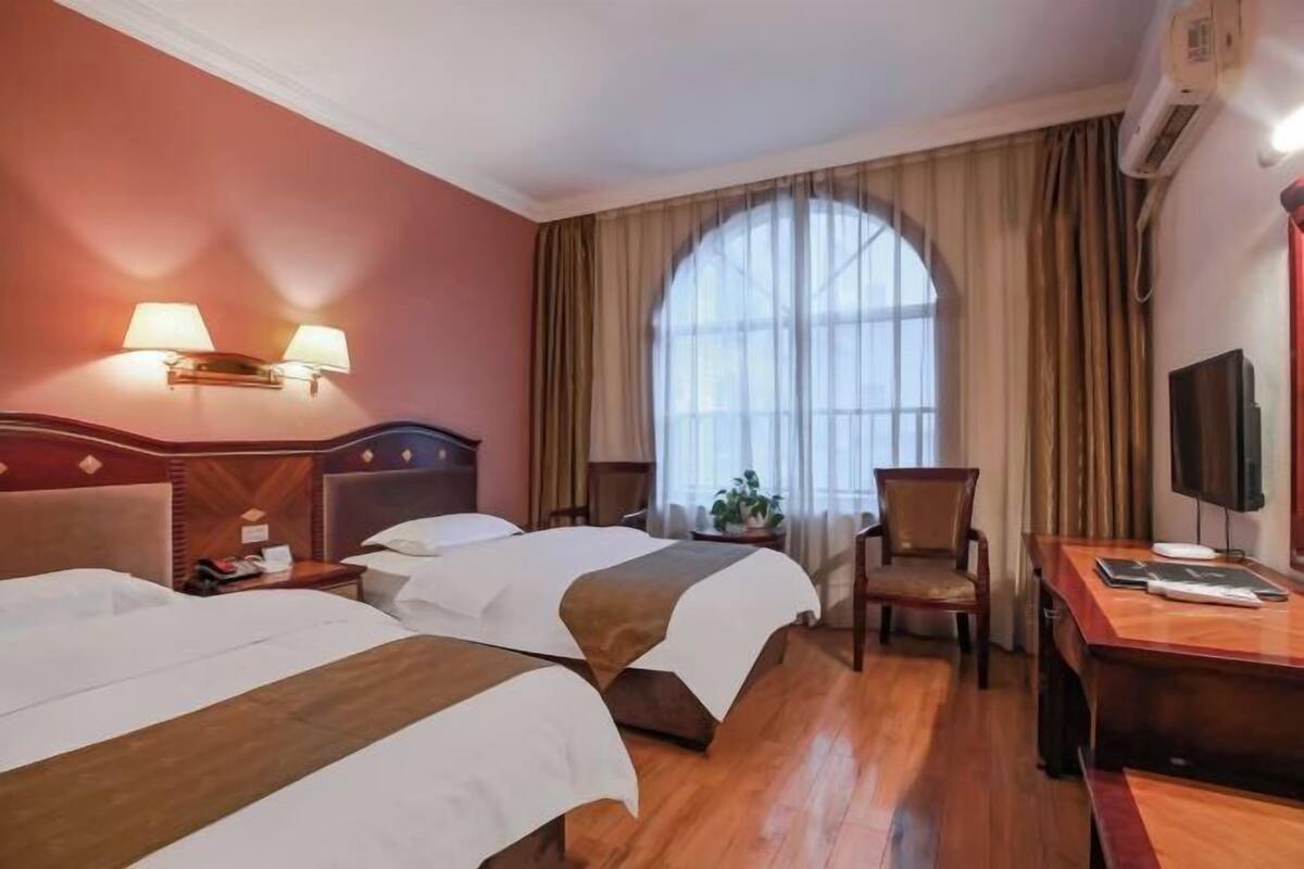 A hotel room with two beds, a desk, a chair, a TV, and a large window with curtains.