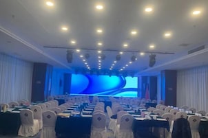 Un salón de conferencias con filas de mesas y sillas, una pantalla grande al frente y un escenario con un telón de fondo azul.