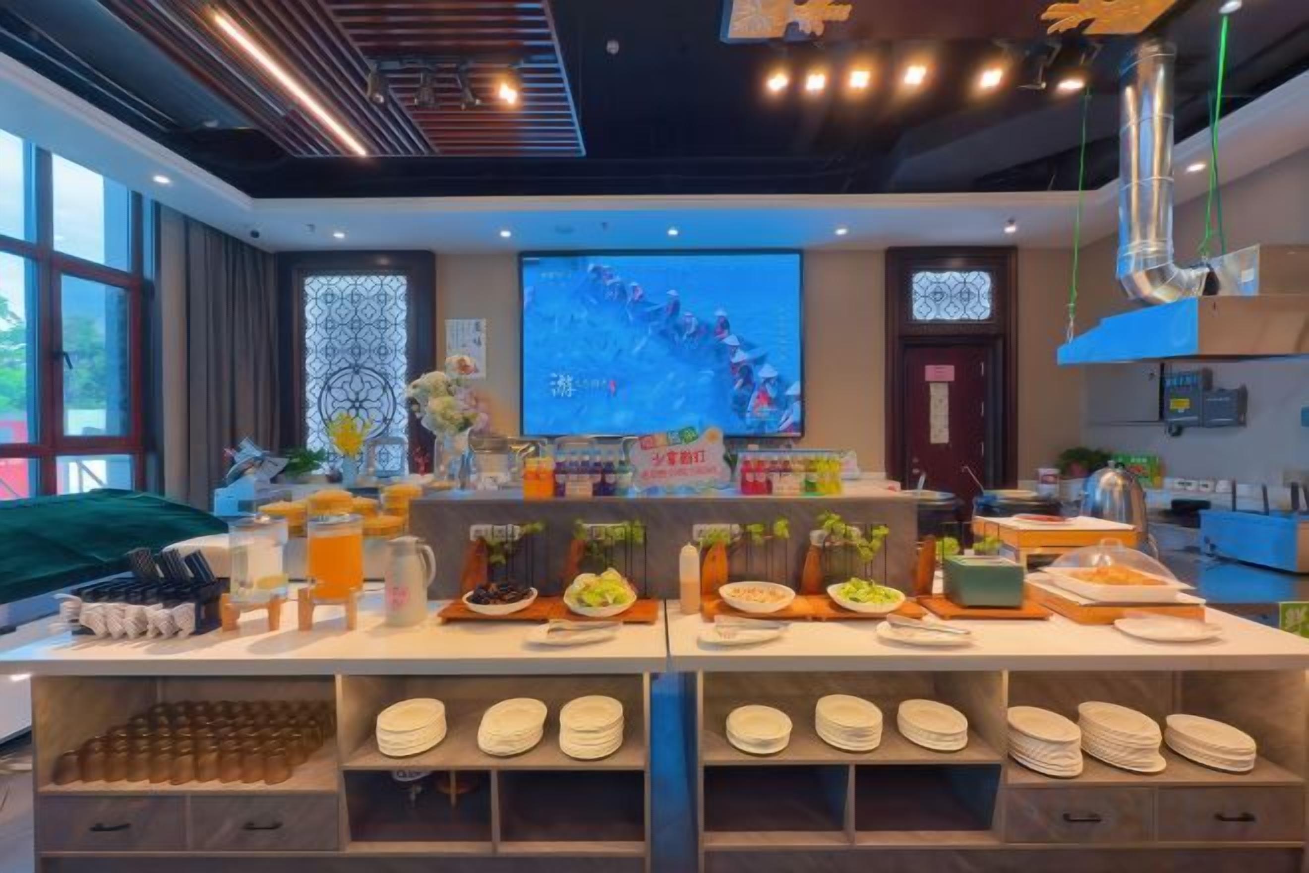 A buffet setup with various food items and beverages, a projection screen in the background, and a kitchen area.